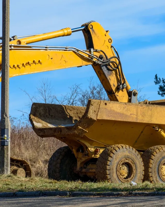 A backhoe dumps a load of backfill into the back of a scratched up dump truck.