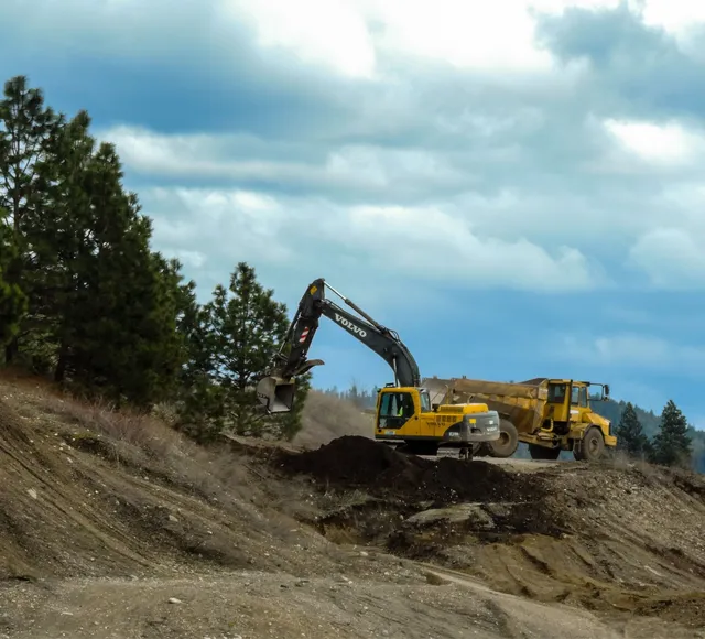 A backhoe removes soil from a hillside, placing it in the back of a large yellow construction dump truck.