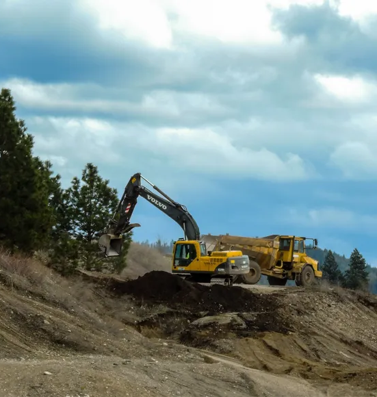A backhoe removes soil from a hillside, placing it in the back of a large yellow construction dump truck.